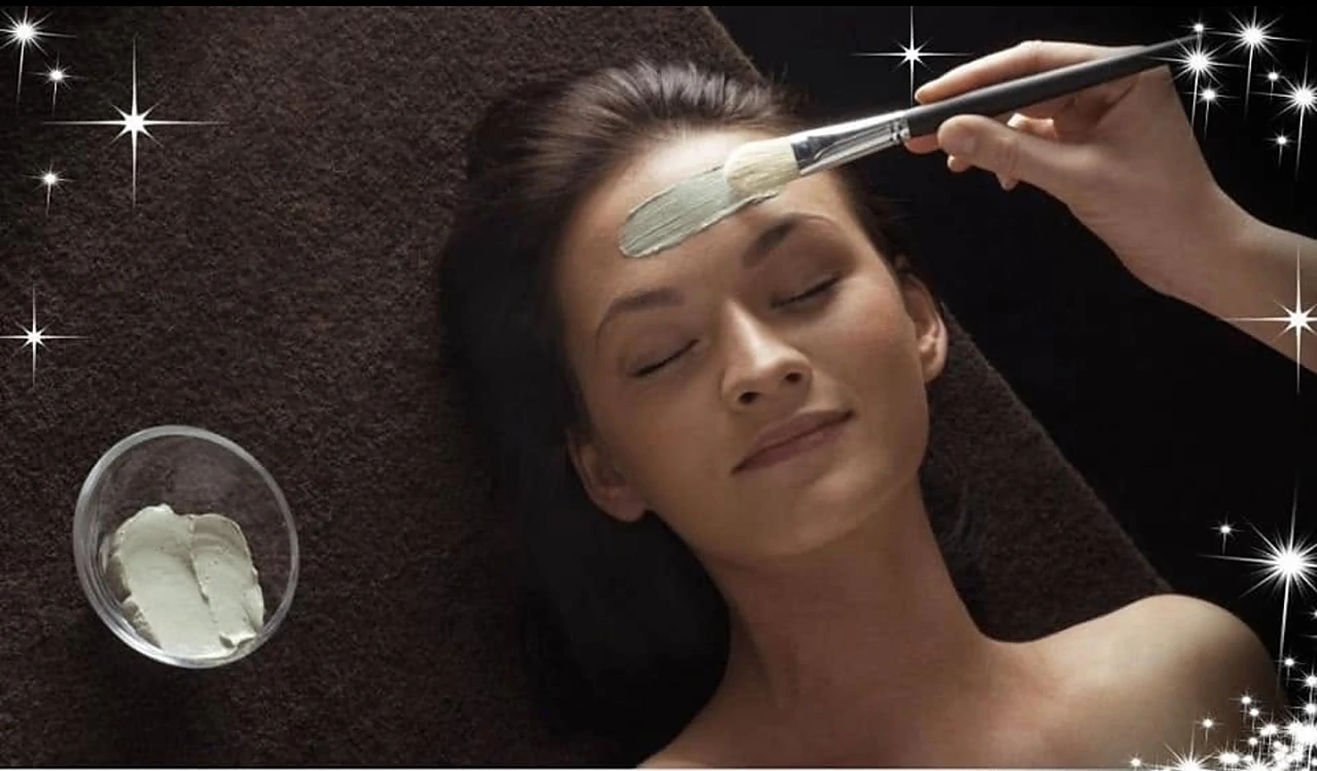 A client relaxing with her eyes closed while a therapist brushes a clay treatment mask across her forehead.